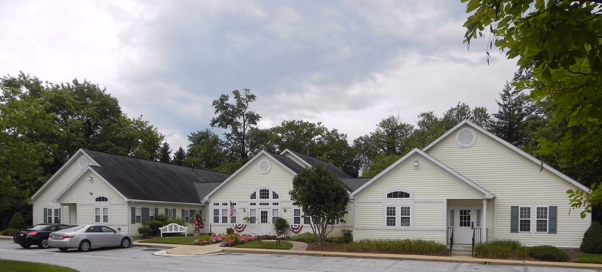 Exterior view of a single-story senior living facility building with cream-colored siding, multiple gabled roofs, and several windows. There are a few parked cars in front, a small garden with flowers, a white bench, and an American flag near the entrance. Trees and greenery surround the building under a cloudy sky.