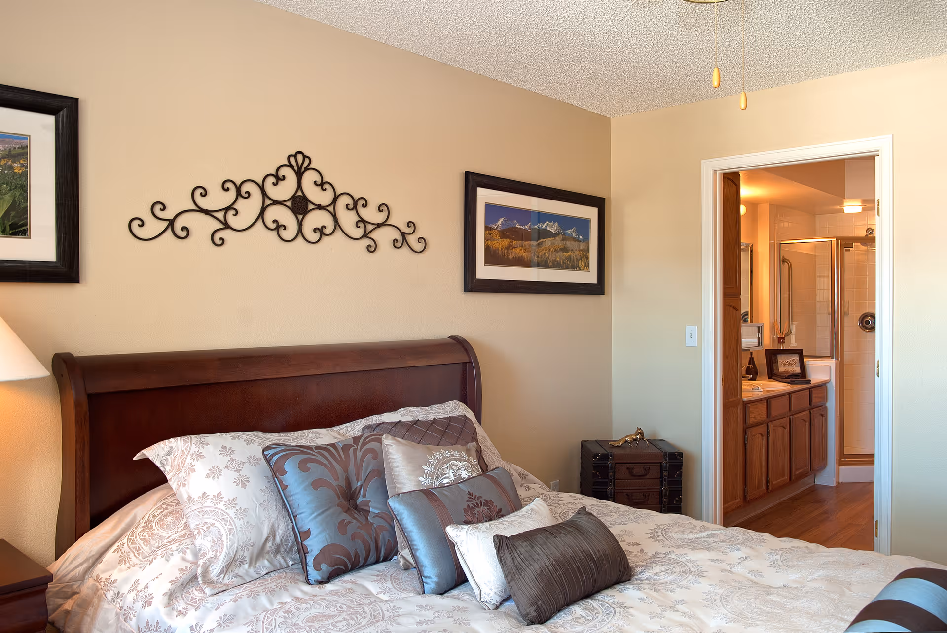 A cozy bedroom with a wooden bed frame and a bed made with patterned bedding and multiple decorative pillows. The walls are beige and decorated with framed landscape pictures and a decorative metal wall art piece. A door leads to an adjoining bathroom with wooden cabinets and a glass shower enclosure.