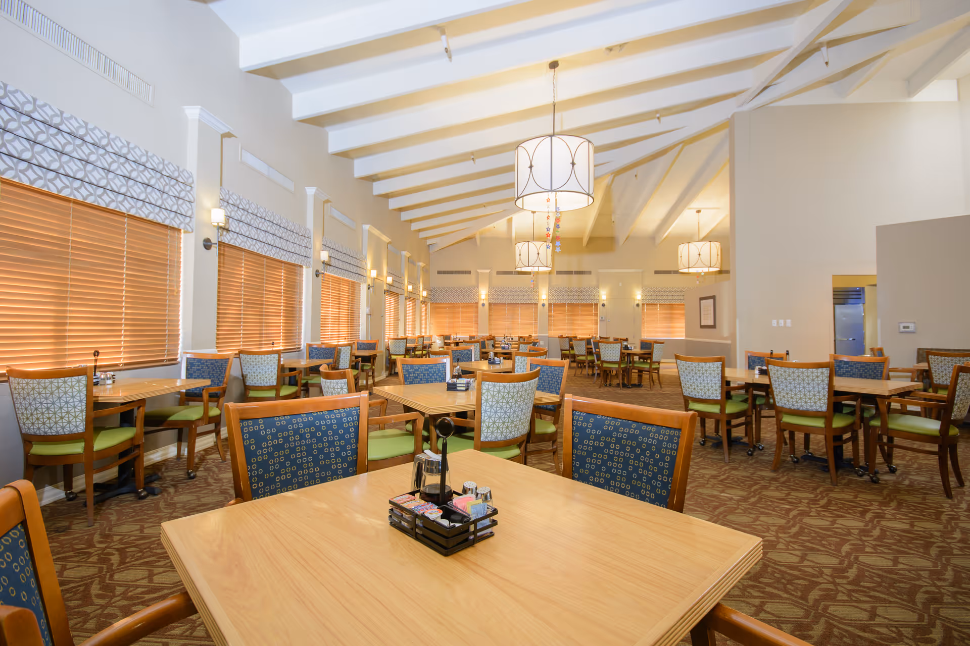 Spacious, well-lit dining room with multiple wooden tables and patterned upholstered chairs under a vaulted ceiling with pendant lights.