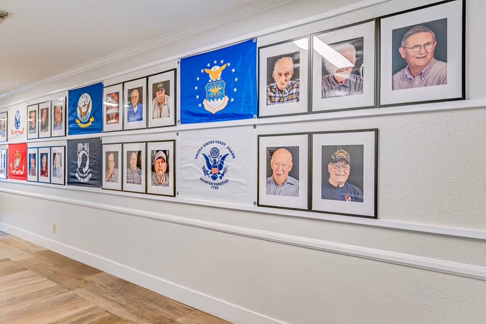 Interior hallway wall with framed portraits of residents and several military service flags displayed.