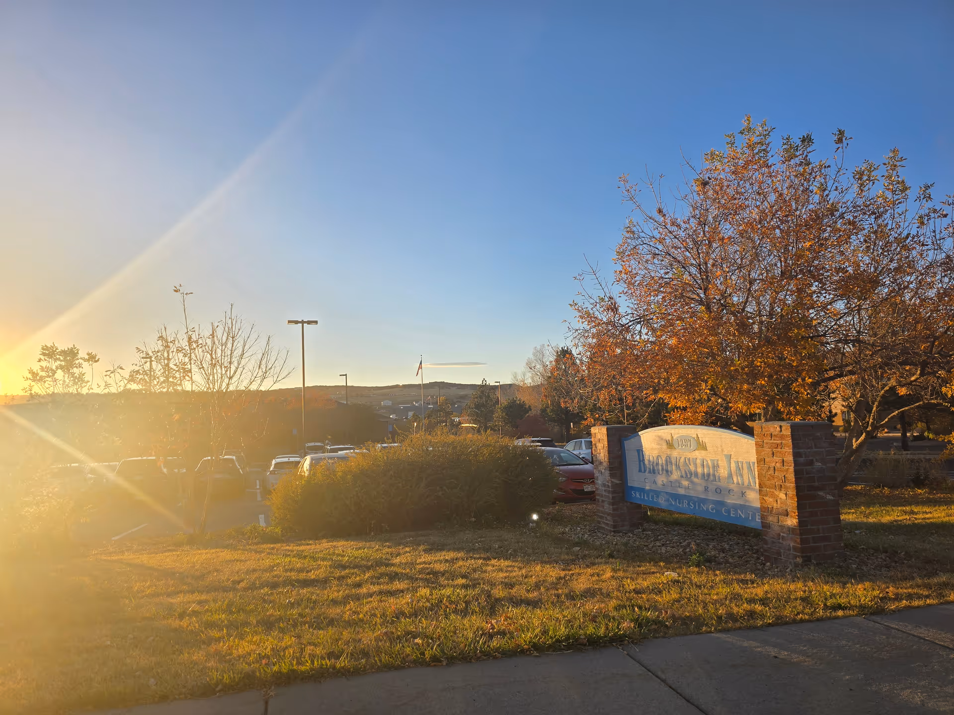 Outdoor view of the Brookside Inn Skilled Nursing Center sign with autumn trees and a parking lot in the background during sunset.