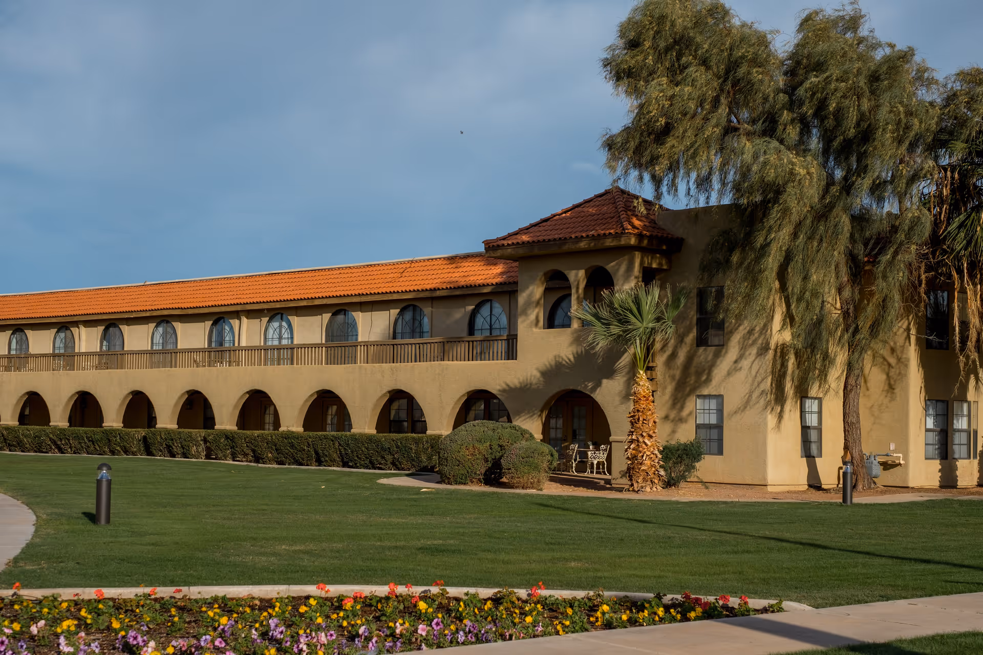 Exterior view of The Garnet of Casa Grande building with beige walls, arched windows and doorways, a red-tiled roof, surrounded by green grass, bushes, palm trees, and a flower bed in the foreground under a clear sky.