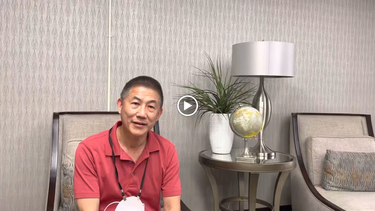A man wearing a red polo shirt sitting in a beige armchair in a room with patterned wallpaper. Next to him is a round side table with a white vase containing a green plant, a small globe, and a silver table lamp. Another beige armchair with a decorative pillow is partially visible on the right.