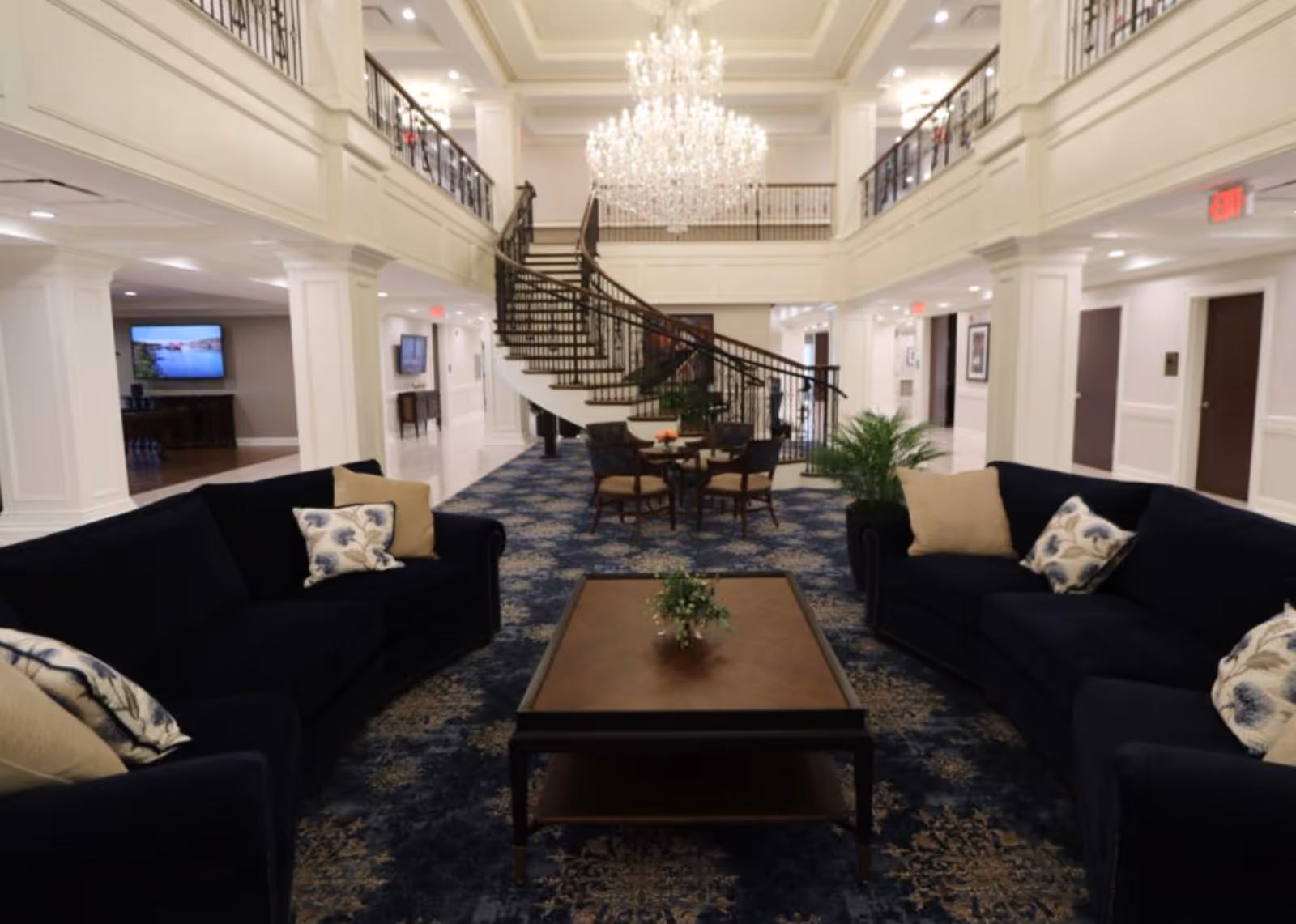 A spacious and elegant senior living facility common area with two dark blue sofas adorned with beige and floral pillows facing a wooden coffee table with a small plant. In the background, there is a grand curved staircase with black railings and a large crystal chandelier hanging from the ceiling. The area is carpeted with a blue patterned rug and has white walls with decorative molding. There are additional seating areas and hallways visible on either side.