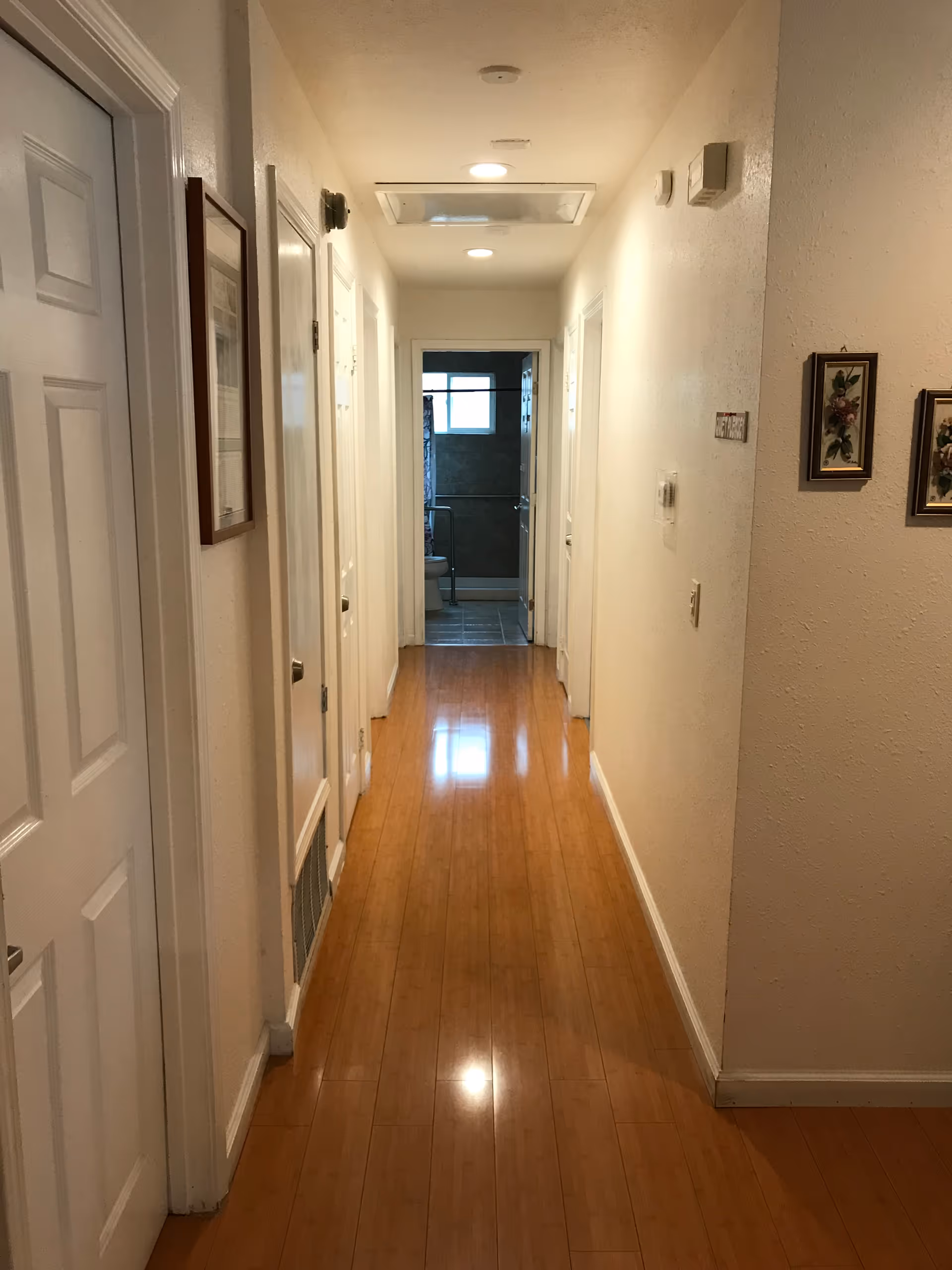 A narrow hallway with wooden flooring and cream-colored walls. Several white doors line both sides of the hallway, and framed pictures hang on the right wall. At the end of the hallway, a bathroom with a window and tiled floor is visible.