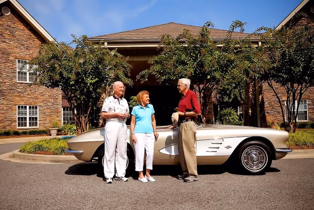 Three elderly people standing and talking in front of a classic silver convertible car parked outside a brick building with trees and landscaping around.