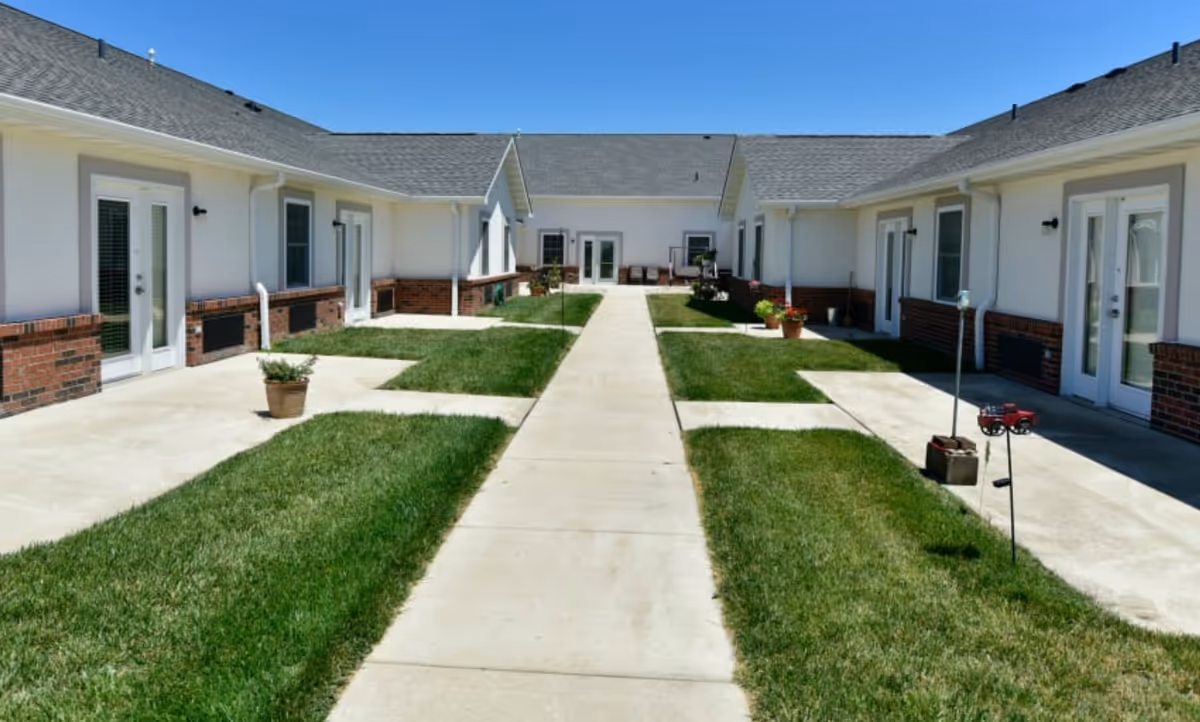 Central paved walkway through a grassy courtyard flanked by single-story residential units with patios and glass doors under a blue sky.