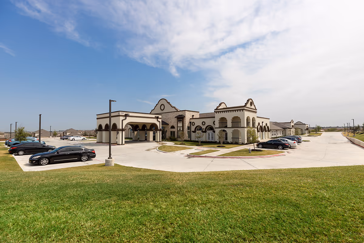 Exterior view of The Grandview of Chisholm Trail senior living facility showing a large building with arched architectural features, a covered entrance, parked cars, and a well-maintained grassy area under a partly cloudy sky.