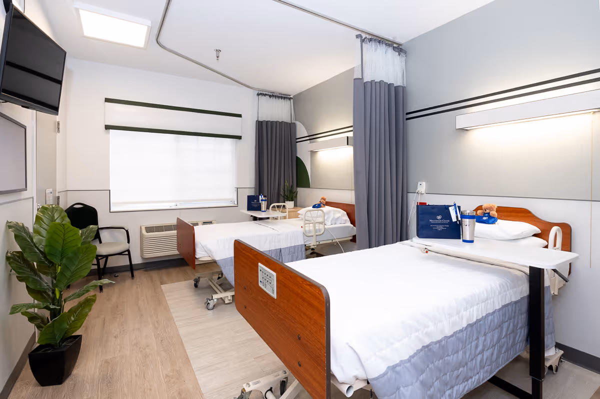 A bright and clean rehabilitation room with two hospital beds separated by a gray privacy curtain. Each bed has white linens and a small overbed table. There is a window with a white blind, a wall-mounted TV, a black chair, and a green potted plant. The room has light wood flooring and soft gray walls with a horizontal stripe detail.