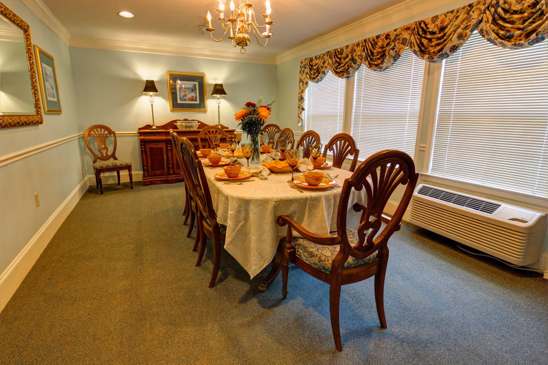 A formal dining room with a long table covered with a white tablecloth, set with plates, bowls, glasses, and napkins. The room has wooden chairs with patterned cushions around the table, a sideboard with two lamps and a framed picture above it, a large mirror on one wall, and windows with blinds and floral valances. There is also a wall-mounted air conditioning unit beneath the windows.
