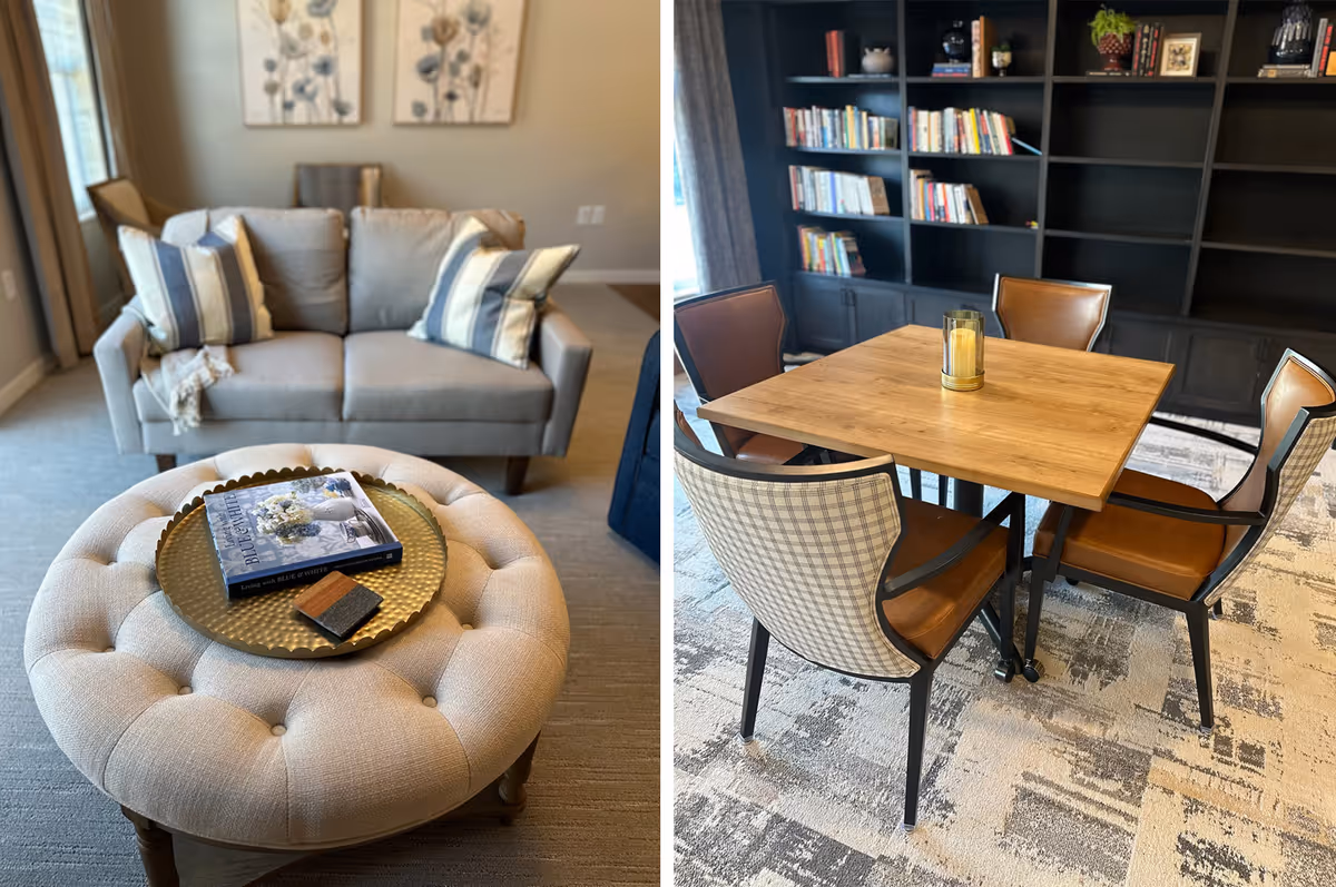 A cozy living area with a beige tufted ottoman topped with a decorative tray holding a book and a small item, positioned in front of a light gray sofa with striped pillows. Adjacent to this is a wooden table surrounded by four chairs with leather seats and patterned backs, set on a patterned carpet with a large dark bookshelf filled with books and decorative items in the background.