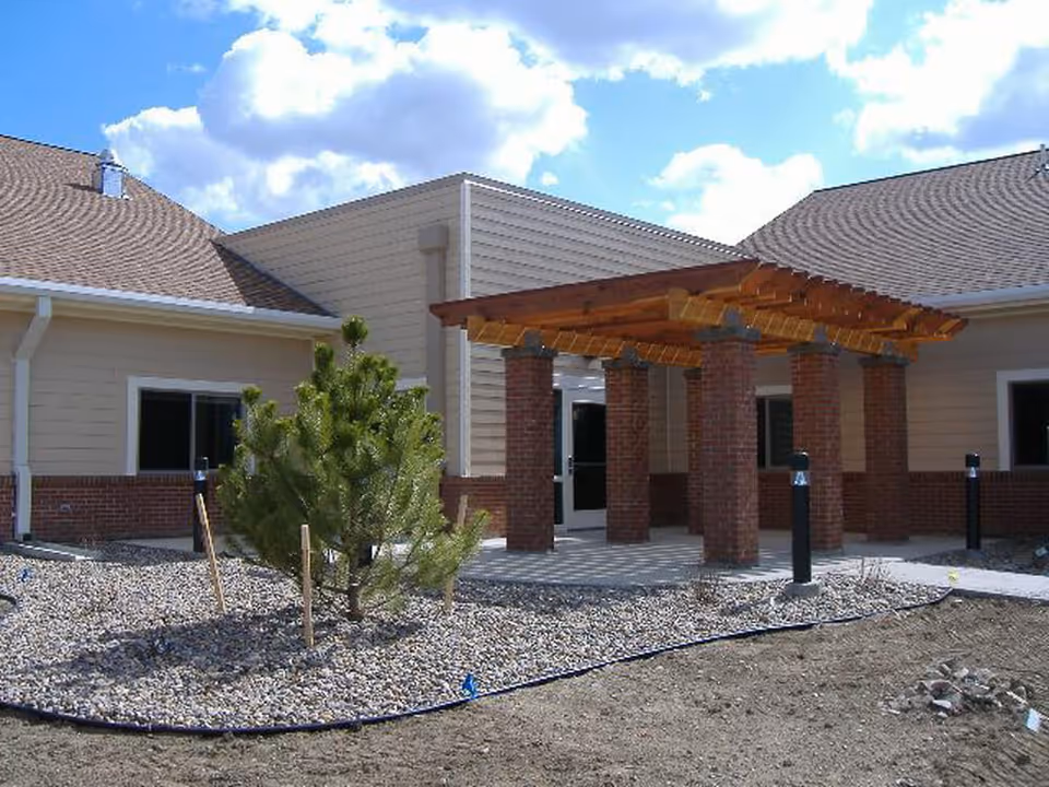 Exterior view of a building with beige siding and brick accents, featuring a wooden pergola supported by brick columns over a paved entrance area. There is a small landscaped area with a young pine tree and gravel in front of the building under a partly cloudy sky.