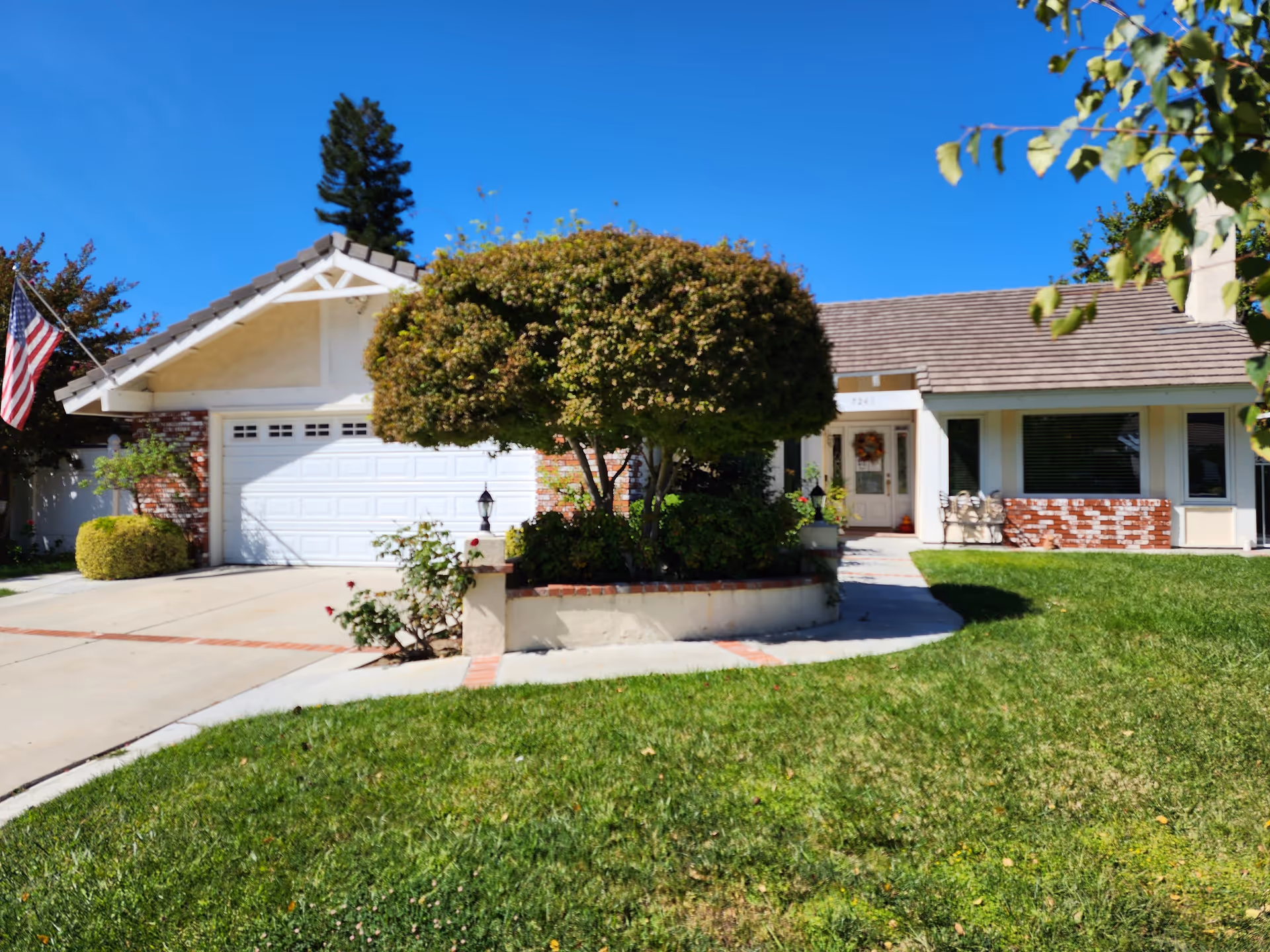 Front exterior of a single-story ranch-style house with a garage, driveway, trimmed tree and a green lawn.