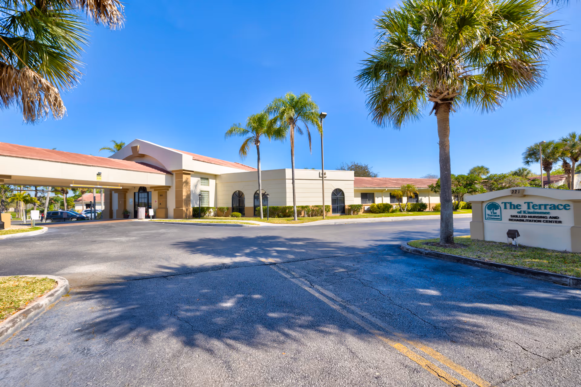 Front entrance and driveway of The Terrace of Kissimmee skilled nursing and rehabilitation center with palm trees and a clear blue sky.