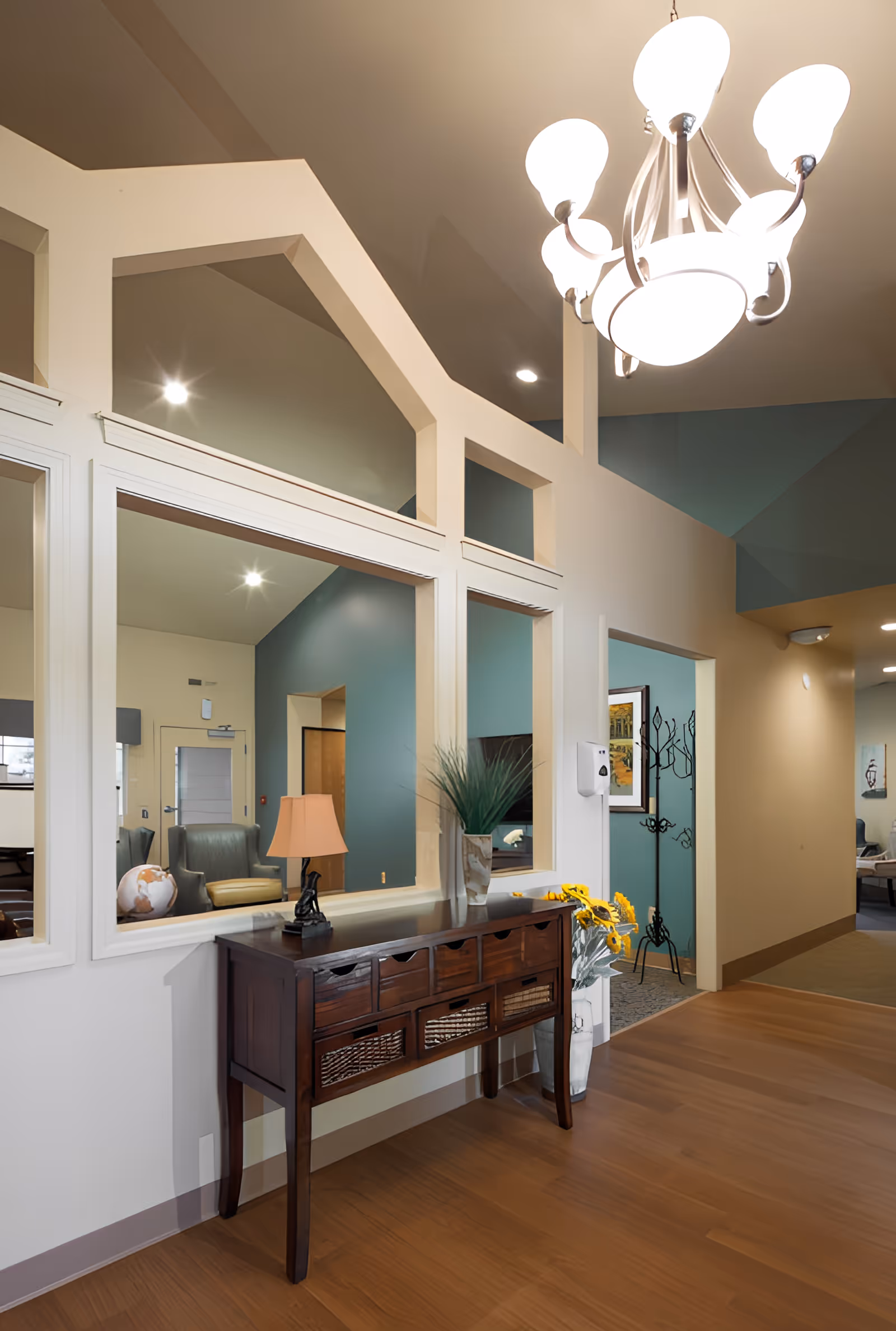 Interior hallway of a senior living facility with wooden flooring, a dark wooden console table with drawers and baskets, a small lamp, a vase with green plants, and a vase with sunflowers. The walls are painted in beige and teal colors, and there are large windows with white frames looking into an adjacent room with armchairs and a globe. A chandelier with multiple lights hangs from the ceiling.