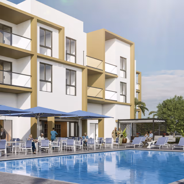 Outdoor pool area at a senior living facility with several people sitting at tables under blue umbrellas and lounging on chairs near the water. The building in the background is modern with white walls and beige accents, featuring balconies and large windows.