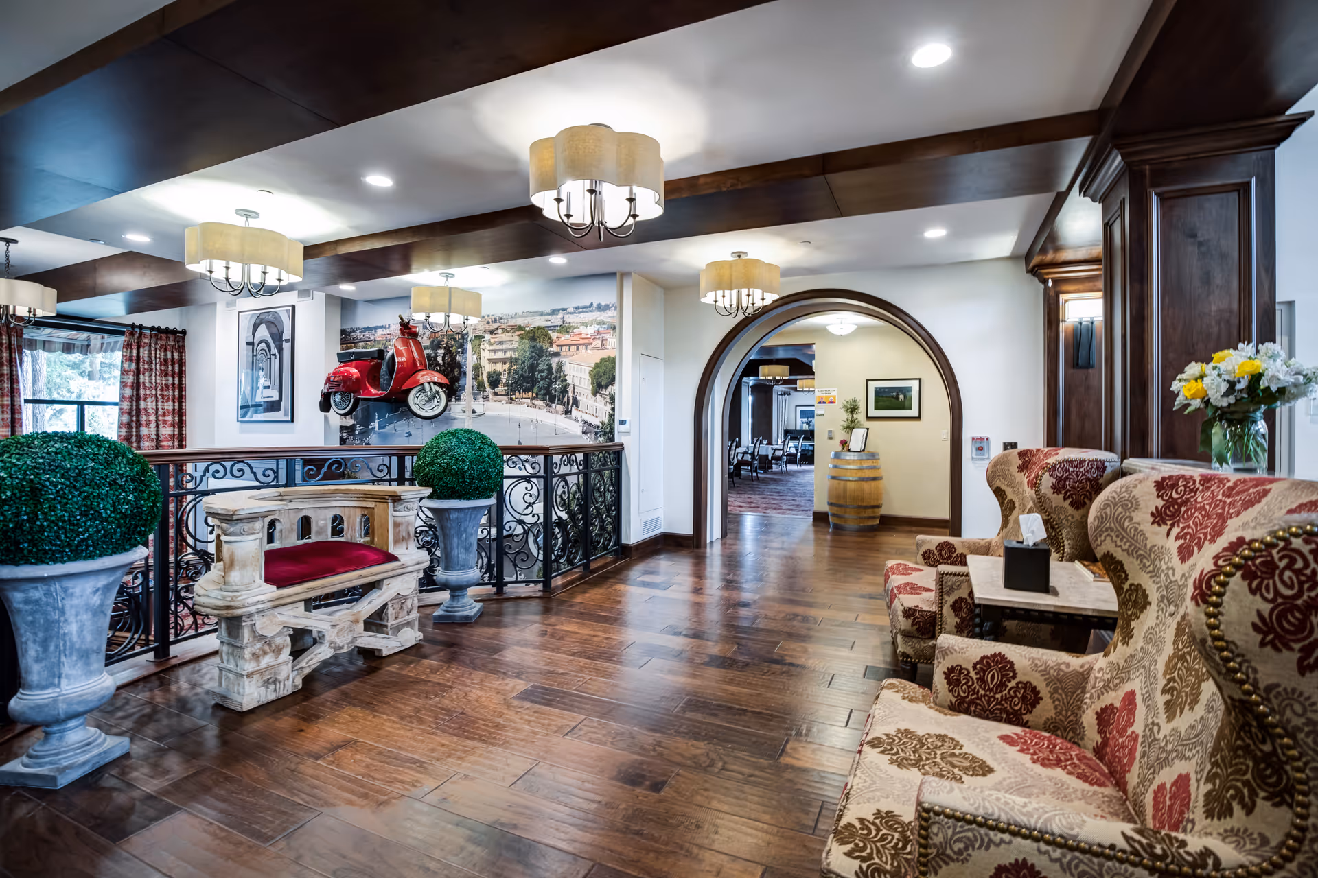 Interior view of a senior living facility lounge area with wooden flooring, decorative ceiling lights, patterned armchairs with a small table, a stone bench with a red cushion, two large potted plants, and a red scooter mounted on the wall above a scenic mural. An arched doorway leads to another room with dining tables and chairs.