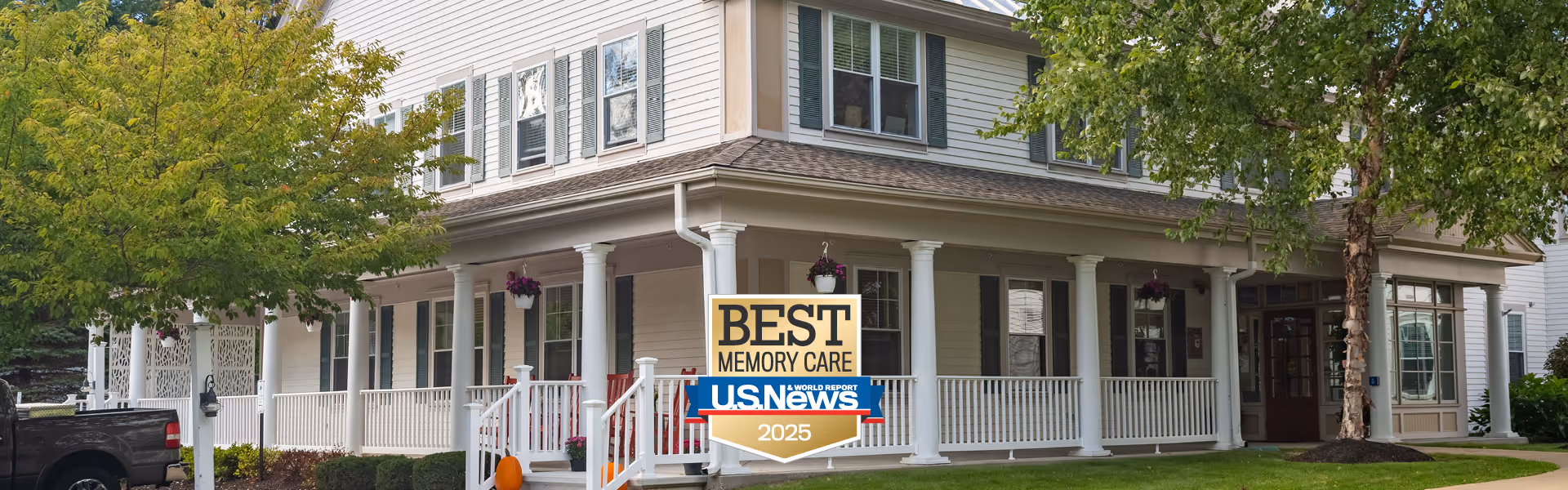 Exterior view of a large two-story senior living facility with white siding, multiple windows, and a wrap-around porch with white railings and columns. There are trees and shrubs around the building, and a black pickup truck is parked nearby. A sign in front of the porch reads 'Best Memory Care U.S. News 2025'.