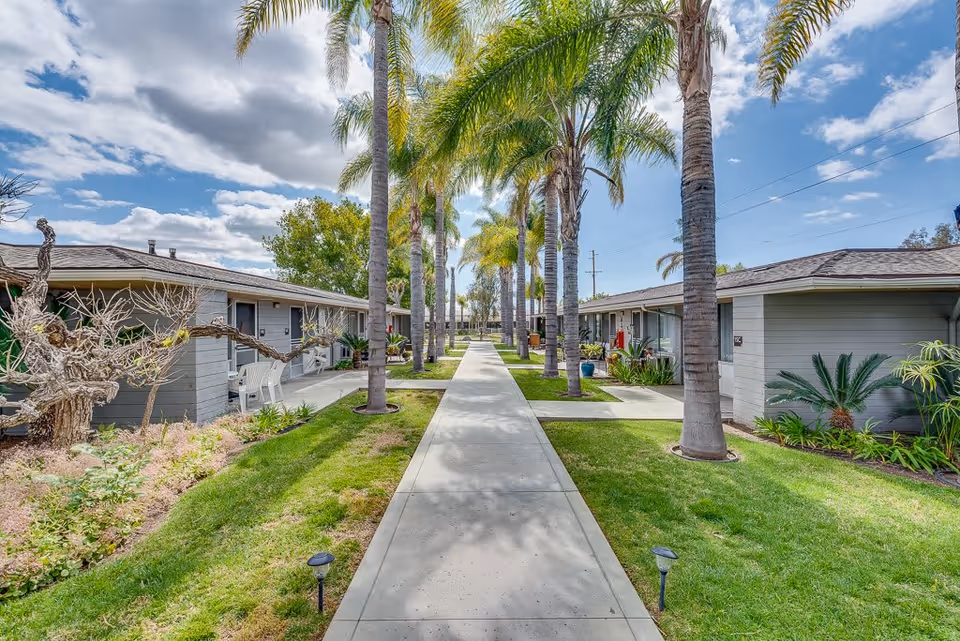A paved walkway lined with tall palm trees runs between two single-story buildings with gray siding and sloped roofs. The area is landscaped with green grass, small plants, and shrubs under a partly cloudy sky.