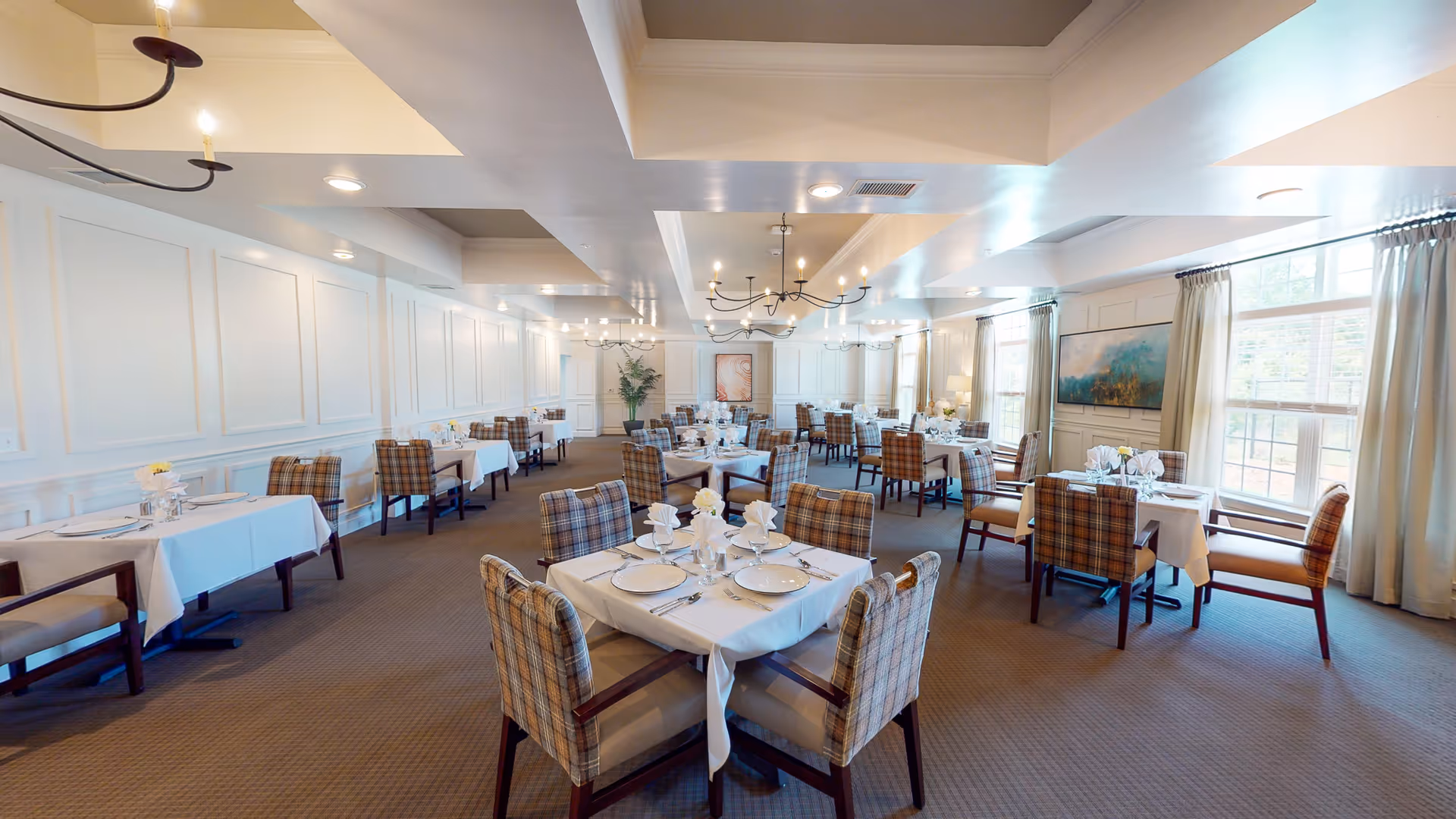 A spacious dining room featuring neatly arranged tables with white tablecloths, elegant place settings, and comfortable plaid chairs. Large windows allow natural light to fill the room, complemented by decorative chandeliers and artwork on the walls.