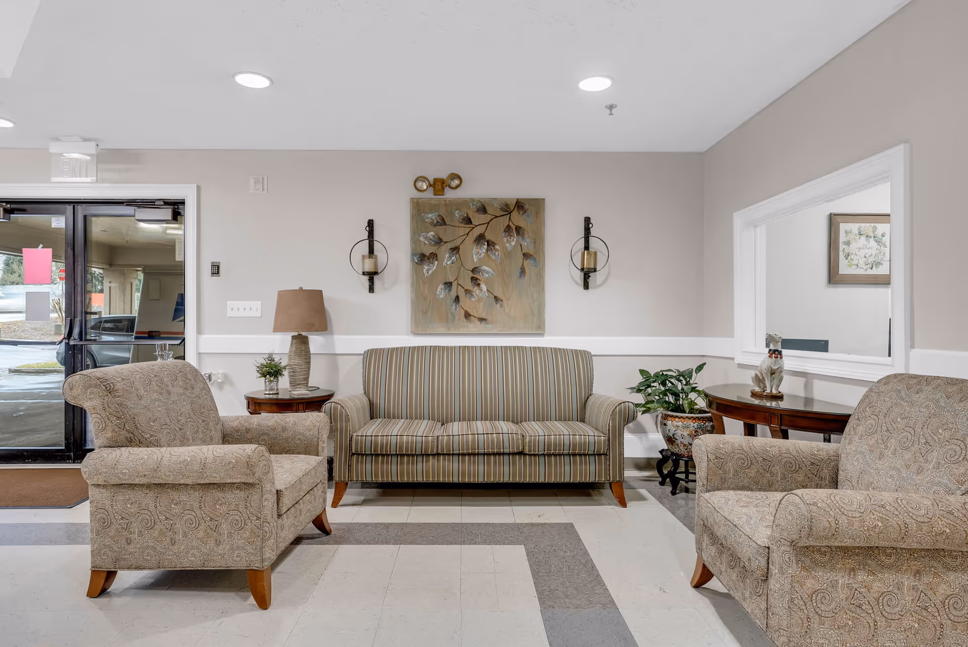 A cozy seating area in a senior living facility with two patterned armchairs and a striped sofa arranged around a small wooden side table with a lamp and a plant. There is a decorative wall art piece with leaves above the sofa, two wall-mounted candle holders, and a large window opening to another room with framed artwork and a small statue on a table. The entrance door with glass panels is visible on the left side.