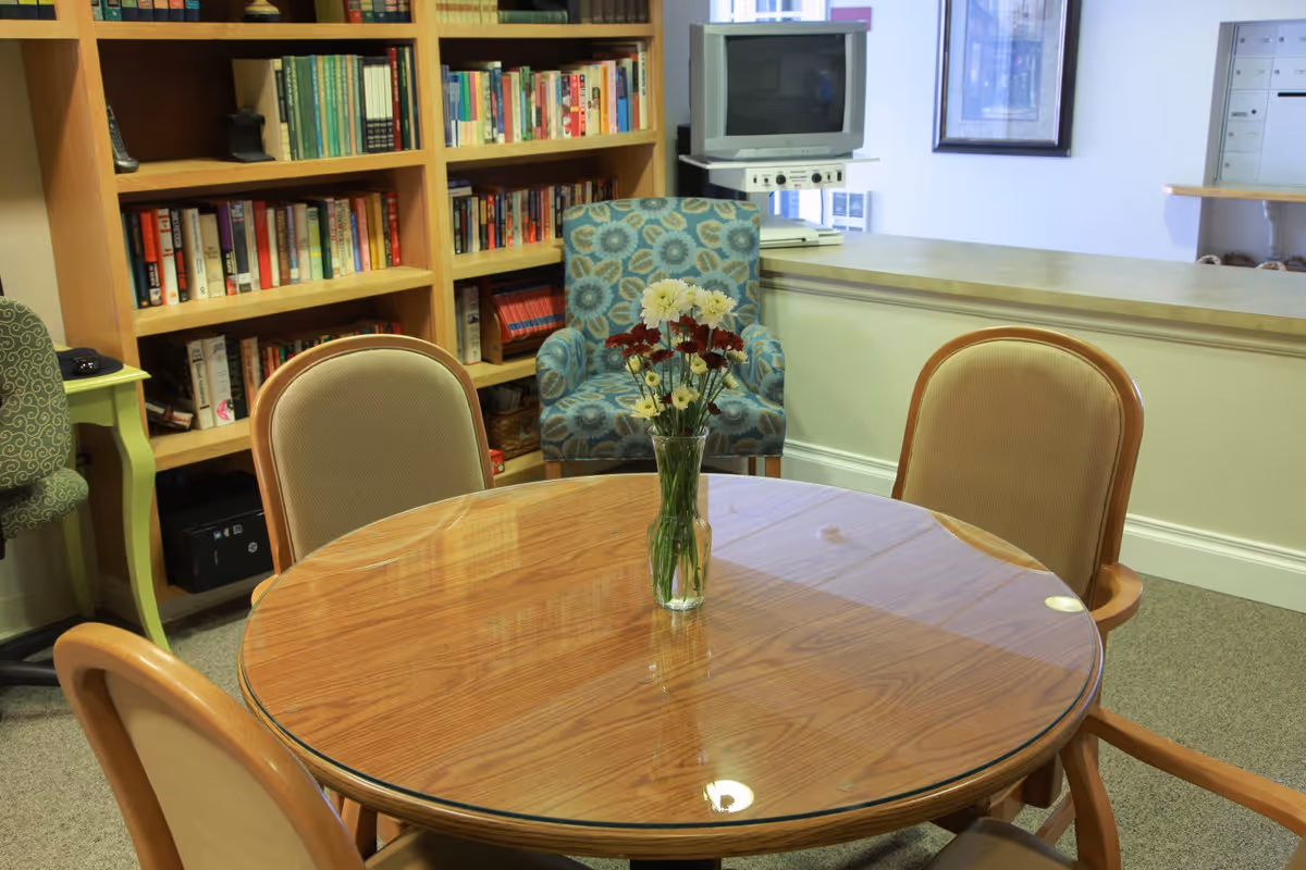 A cozy interior room featuring a round wooden table with a glass top and a vase of flowers in the center. Four cushioned chairs surround the table. Behind the table is a bookshelf filled with books, a patterned armchair, and a small television on a stand. The room has a carpeted floor and light-colored walls.