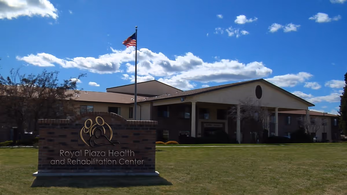Exterior view of the Royal Plaza Health and Rehabilitation Center building with a brick sign in front and an American flag on a flagpole against a blue sky with scattered clouds.