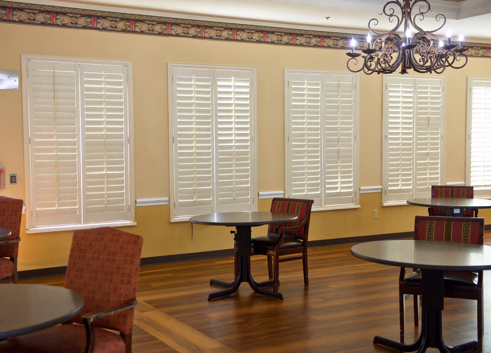 Bright dining room with round tables, upholstered chairs, shuttered windows, and a chandelier.