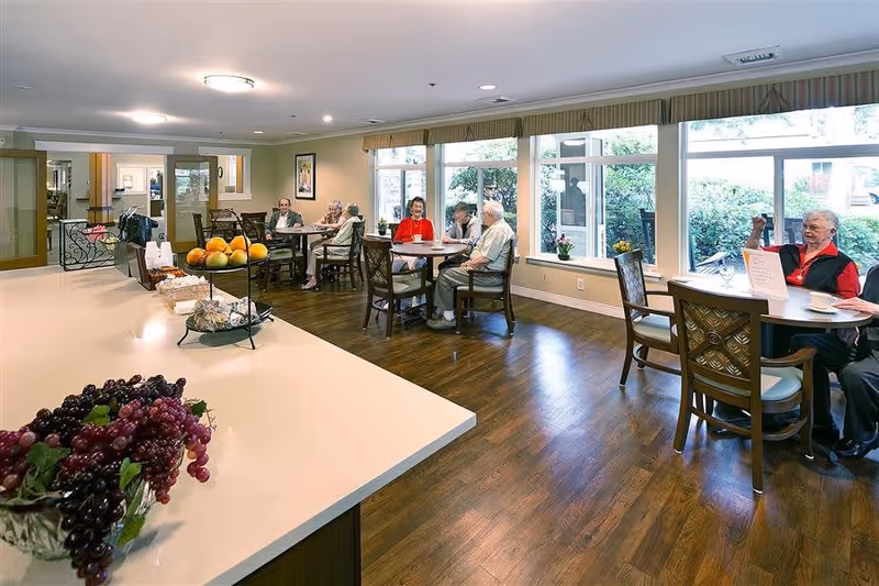 A bright dining area in a senior living facility with several elderly residents sitting at tables, enjoying conversation and drinks. The room features large windows letting in natural light, wooden flooring, and a countertop with fruit and snacks in the foreground.