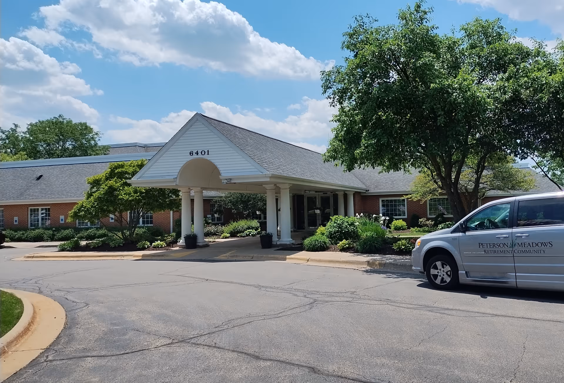 Exterior view of Peterson Meadows retirement community building with a covered entrance, surrounded by trees and landscaping. A silver van with Peterson Meadows Retirement Community logo is parked near the entrance under a partly cloudy sky.