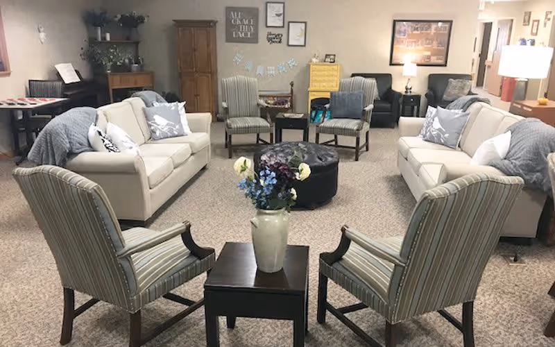 A cozy living room area with two beige sofas, four striped armchairs, a black round ottoman, and a small table with a vase of flowers in the center. The room has carpeted flooring, a piano in the corner, and various wall decorations including framed pictures and signs.