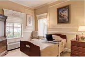 A cozy bedroom with a single wooden bed covered with a beige quilt. A laptop is placed on a tray on the bed. There is a wooden nightstand with a lamp and some decorative items on the right side of the bed. The room has beige walls with white trim, a framed painting above the bed, a window with white blinds and a cushioned chair next to it. A wooden cabinet is partially visible on the left side.