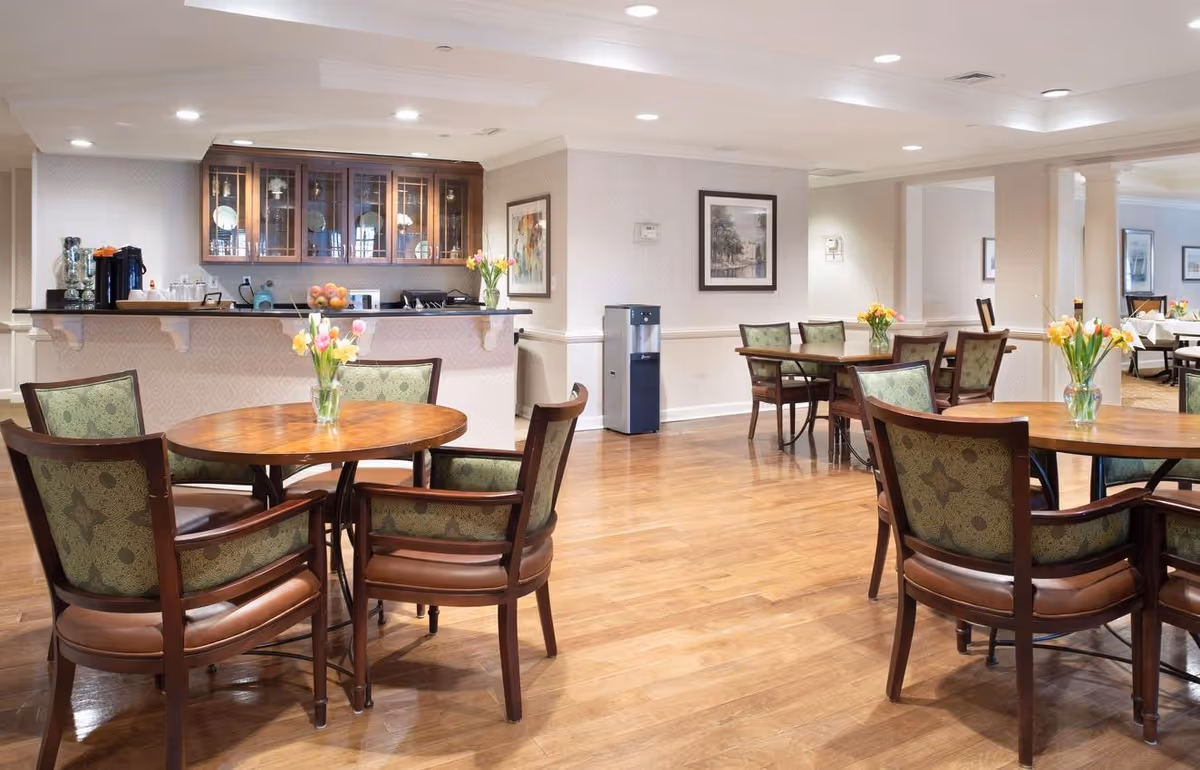 Spacious dining room with round wooden tables, upholstered chairs, vases of flowers, and a serving counter with cabinets in the back.