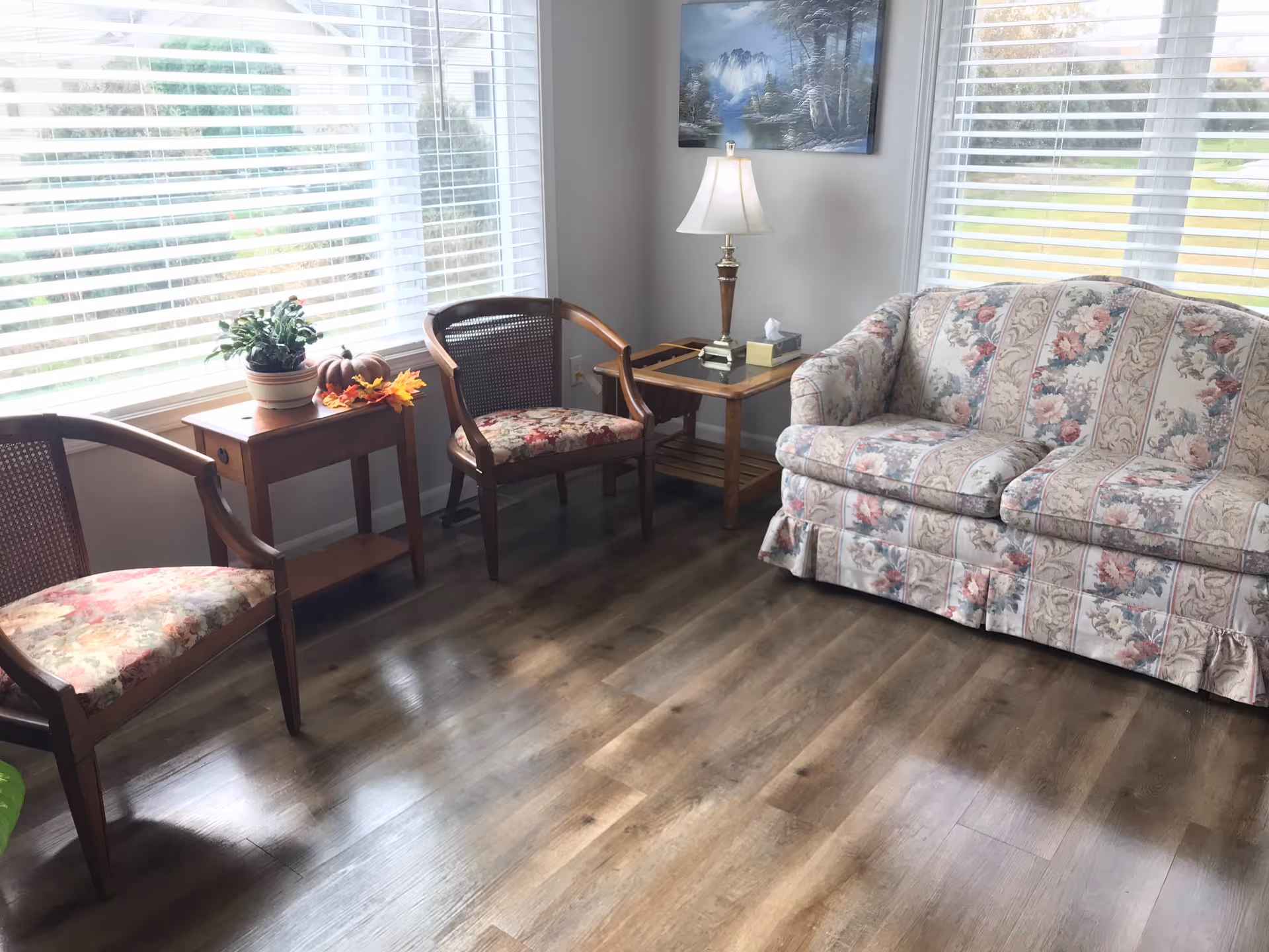 Sunlit living room with a floral sofa, two patterned chairs, wooden side tables, a lamp, and large windows with blinds.