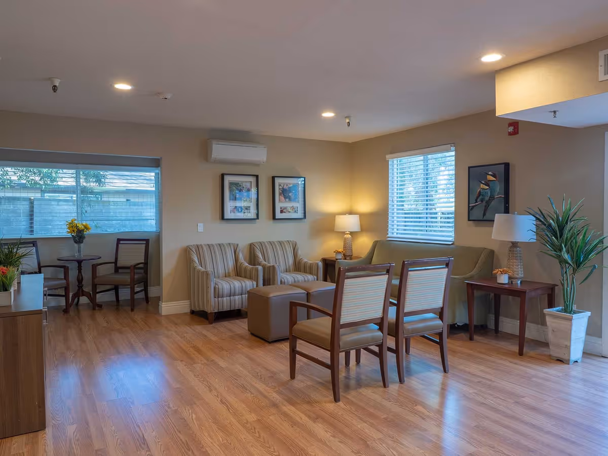 A cozy living room area in an assisted living facility featuring two striped armchairs, two wooden chairs with cushions, a green sofa, two side tables with lamps, framed artwork on the walls, a potted plant, and large windows with blinds allowing natural light to enter.