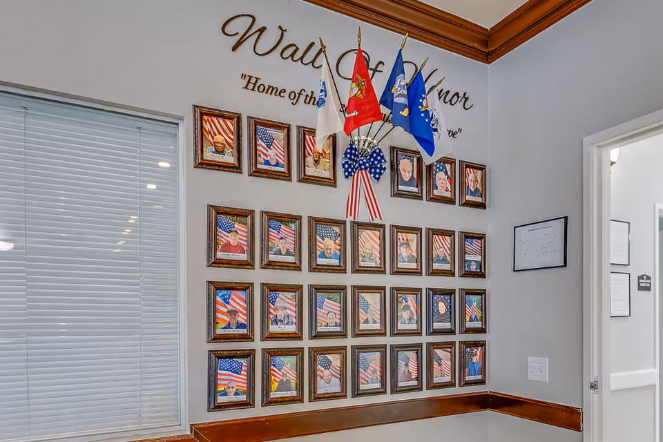 Wall of Honor display featuring framed photographs of veterans with American flags in the background, several military service flags, and a patriotic ribbon, located in an interior hallway of a senior living facility.