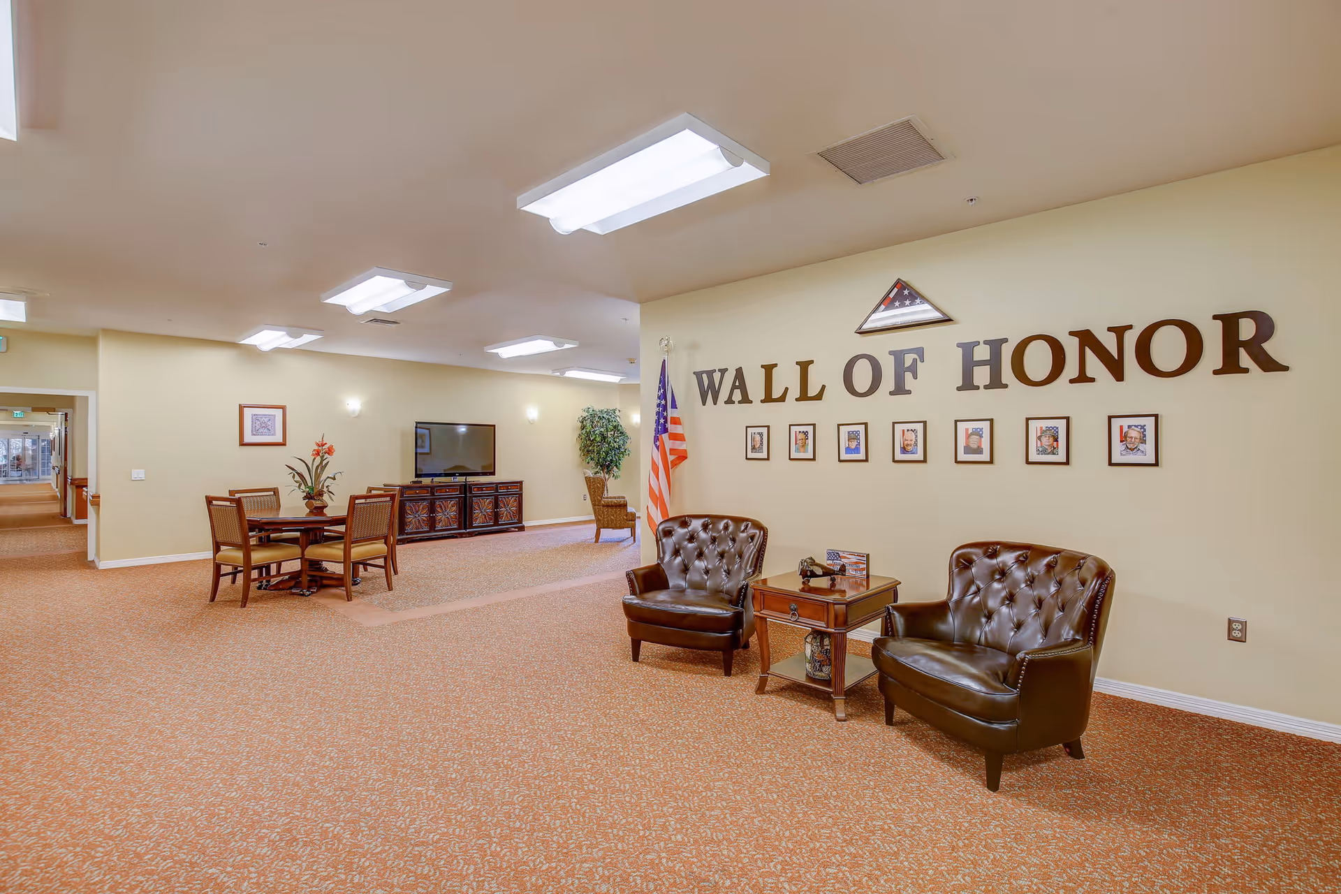Interior view of a senior living facility lounge area with a 'Wall of Honor' display featuring framed photos and a folded American flag. Two brown leather armchairs flank a wooden side table with decorative items. In the background, there is a table with four chairs, a large TV on a cabinet, and an American flag next to a potted plant. The walls are light yellow and the carpet is patterned in warm tones.
