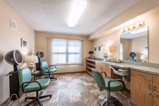 Interior view of a salon room with two green salon chairs and hair dryers on the left side, a large mirror with a countertop and another green salon chair on the right side, beige walls, a window with vertical blinds, and tiled flooring.