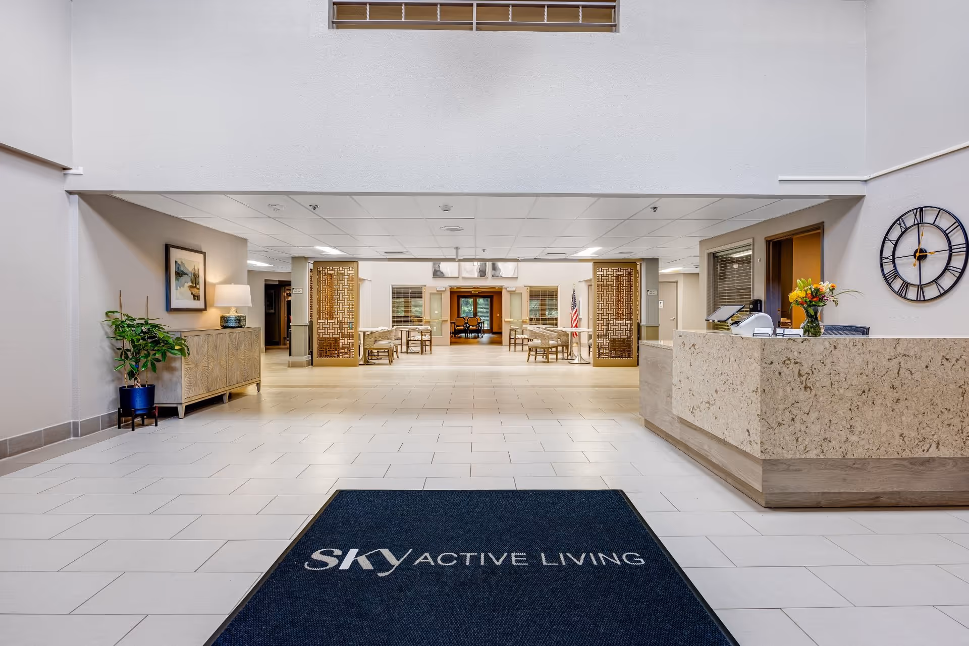 Spacious, well-lit senior living lobby with a reception desk on the right, seating area ahead, and a floor mat reading 'SKY ACTIVE LIVING'.