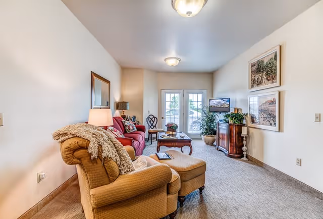 A cozy living room with a beige armchair and matching ottoman in the foreground, a red sofa with pillows, a wooden coffee table, a side table with a lamp, a TV on a wooden cabinet, two framed pictures on the wall, and double glass doors leading outside.