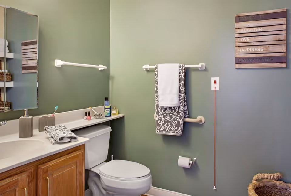 A bathroom with green walls featuring a white toilet, a wooden cabinet with a white countertop, a mirror, and various toiletries. A towel rack holds two towels, and a decorative wooden sign with the words 'Family, Laughter, Love, Happiness, Everything' hangs on the wall. There is also a grab bar and a pull cord near the toilet.