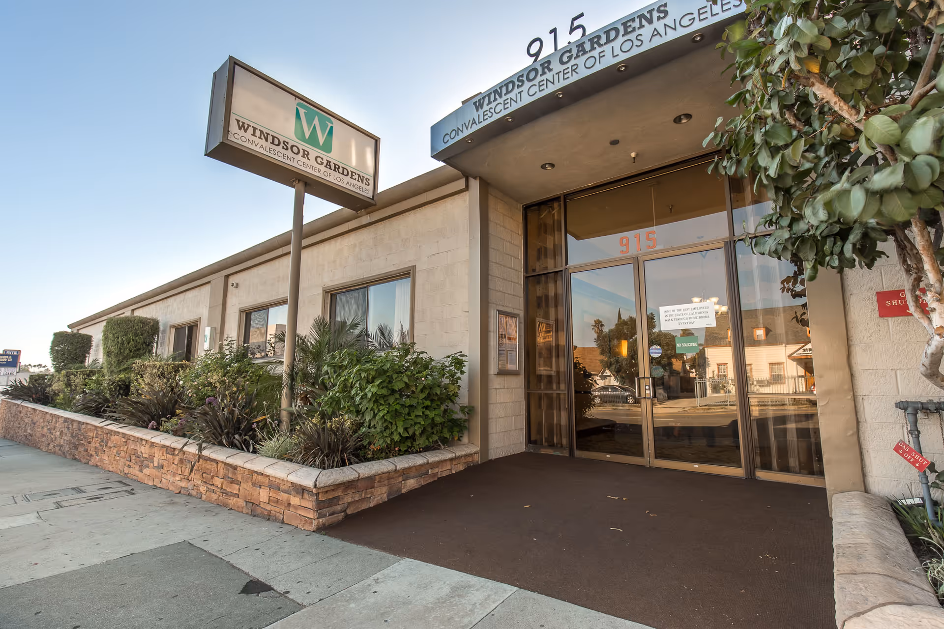 Exterior view of Windsor Gardens Convalescent Center of Los Angeles showing the entrance with glass doors, a sign with the facility name, and surrounding greenery along the sidewalk.