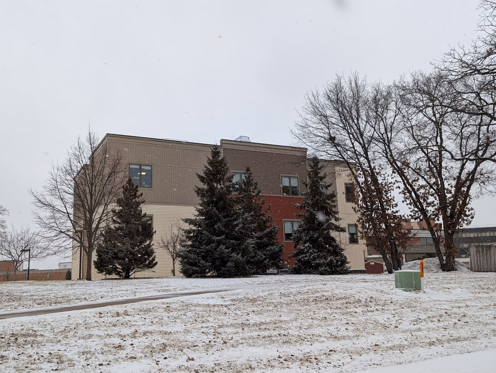 Exterior view of a multi-story building with beige, brown, and red siding partially obscured by evergreen and leafless trees. Snow covers the ground and is falling from the overcast sky.