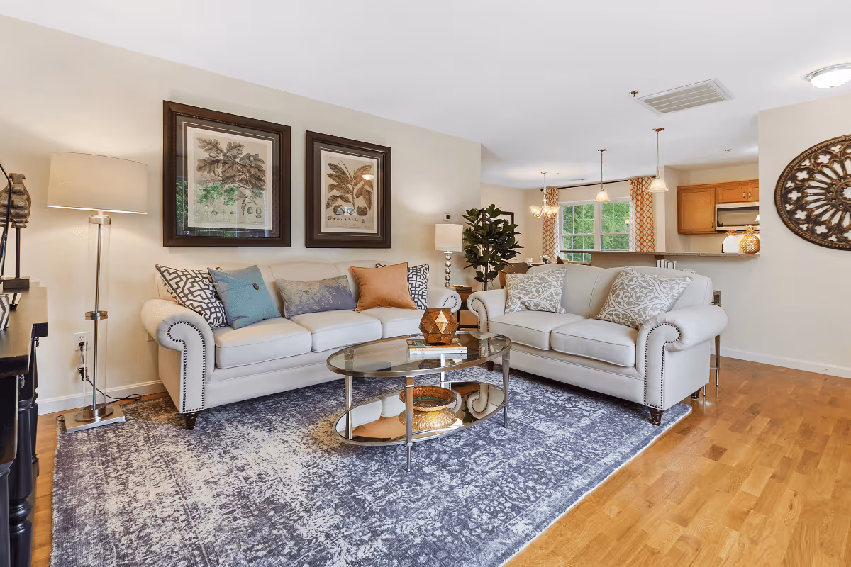 A bright and cozy living room with two beige sofas adorned with decorative pillows, a glass coffee table with books and decor items, a floor lamp, framed botanical artwork on the wall, a patterned area rug, and a view into a dining area and kitchen with wooden floors and light-colored walls.
