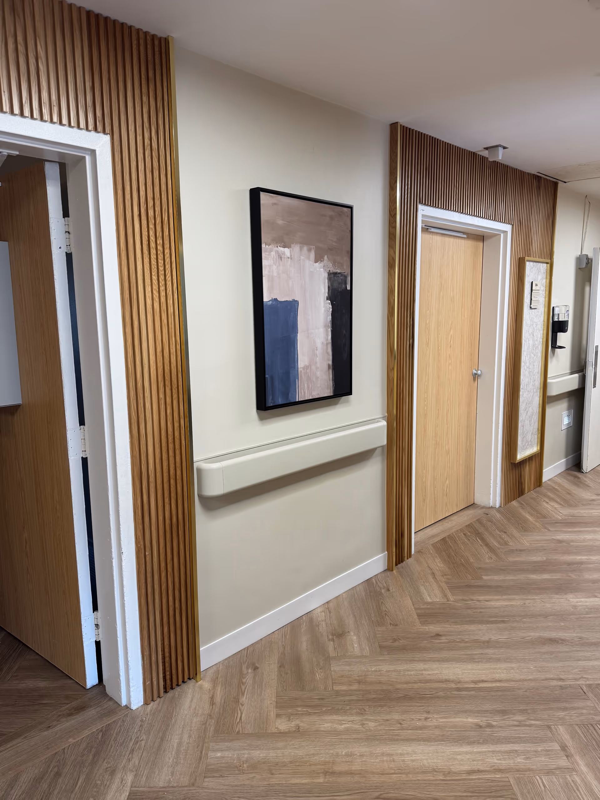 Interior hallway of a facility with light wood flooring arranged in a herringbone pattern, beige walls with vertical wooden panel accents, a handrail along the wall, two closed wooden doors, and abstract artwork hanging on the wall.