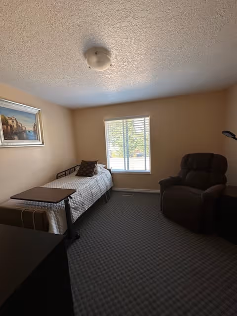 A small bedroom with a single bed covered with a white quilt and a decorative pillow. There is a brown recliner chair next to a window with white blinds. A framed picture hangs on the beige wall above the bed. The floor is carpeted with a patterned design.