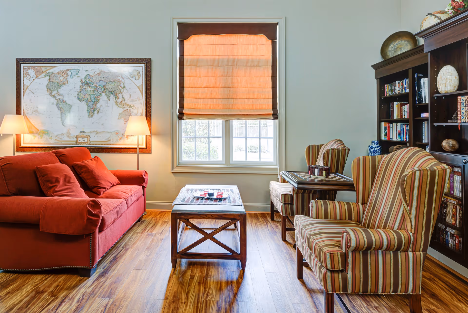 A cozy living room with a red sofa on the left, two striped armchairs on the right, and a wooden coffee table in the center. Behind the sofa is a large framed world map and two floor lamps. A window with an orange Roman shade is centered on the back wall. To the right, there is a dark wooden bookshelf filled with books and decorative items. The floor is wooden with a warm finish.