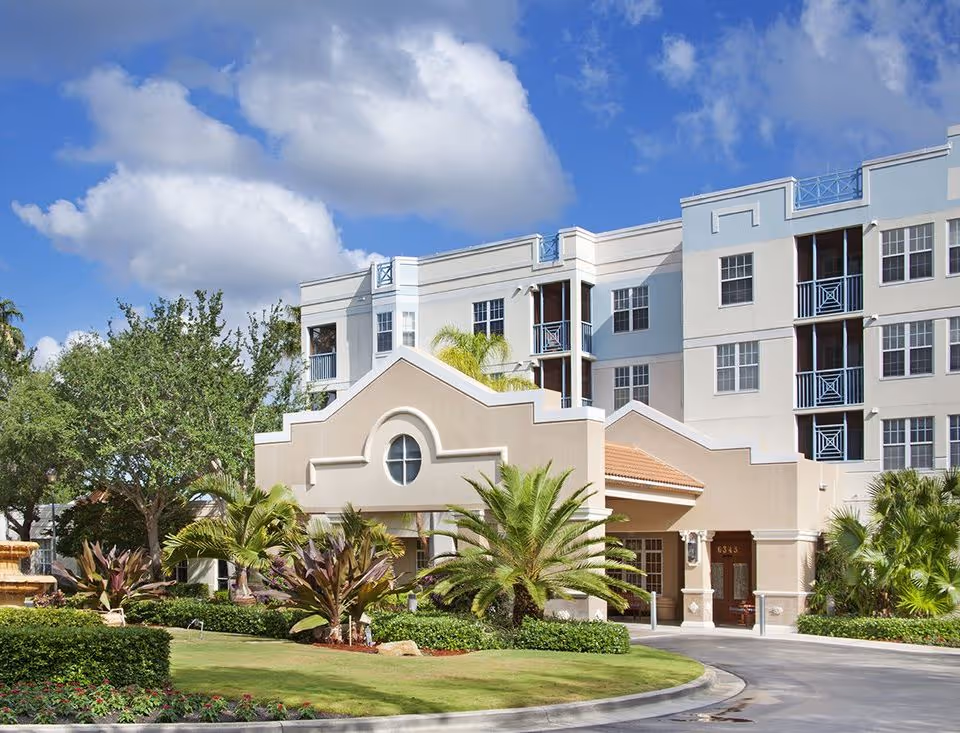 Exterior view of Stratford Court of Boca Pointe, a multi-story senior living facility with beige and light blue walls, surrounded by palm trees, bushes, and a well-maintained lawn under a partly cloudy blue sky.