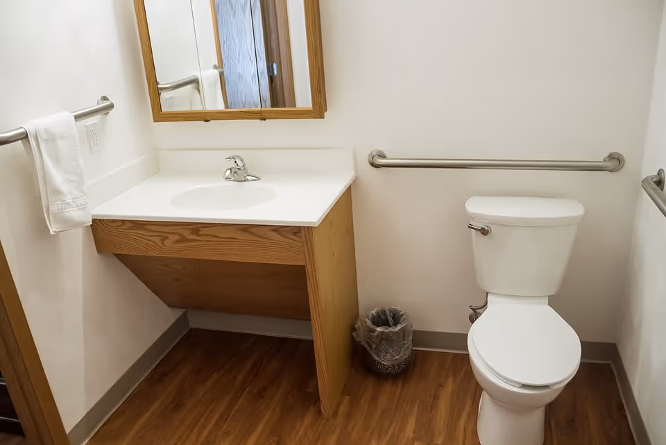 A clean and simple bathroom with a white toilet, a wooden vanity with a white countertop and sink, a mirror above the sink, a towel hanging on a metal bar to the left, and metal grab bars on the walls near the toilet and sink. The floor has a wood-like finish.