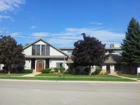 Exterior front view of a two-story residential building with a peaked roof, large windows, and a wooden front door. The building is surrounded by green lawns, bushes, and several trees under a blue sky with some clouds.